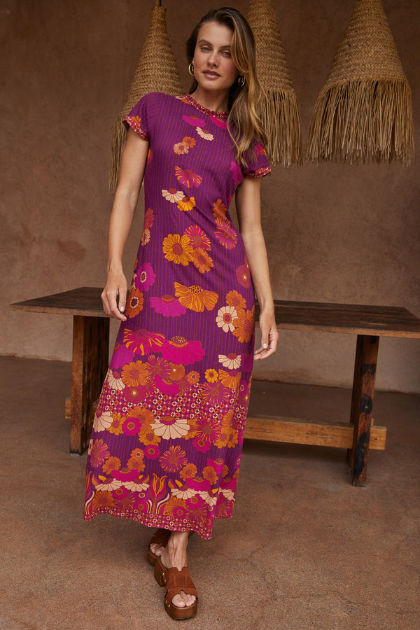 Woman wearing a purple floral dress standing in a rustic setting with wooden bench and straw hats.
