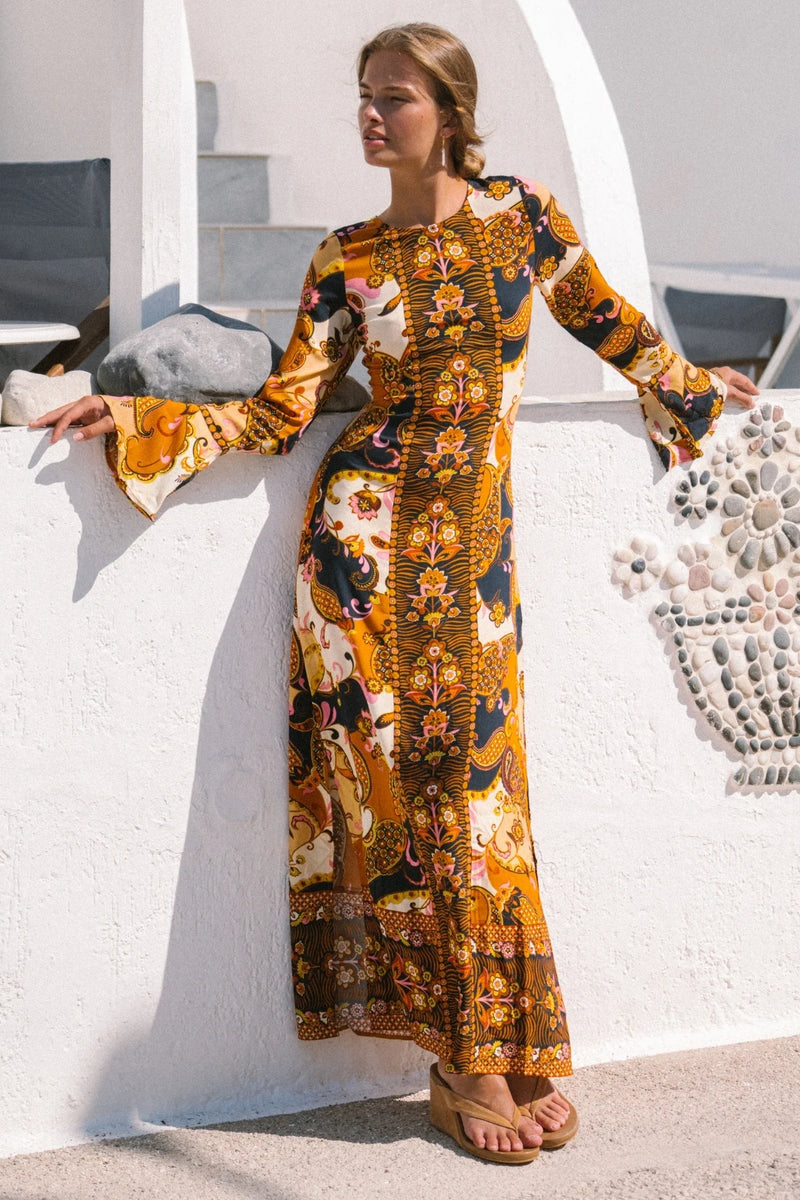 Woman wearing a long, patterned dress standing against a white wall.