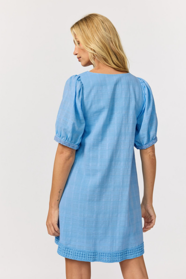 A woman shown from the back wearing a light blue puff-sleeve cotton babydoll mini dress with a crochet trim hem, against a light gray studio background.