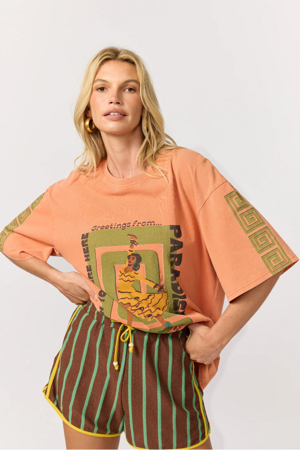 A woman wearing an oversized peach graphic T-shirt with brown and green striped terry towelling shorts with yellow binding, against a light gray studio background.