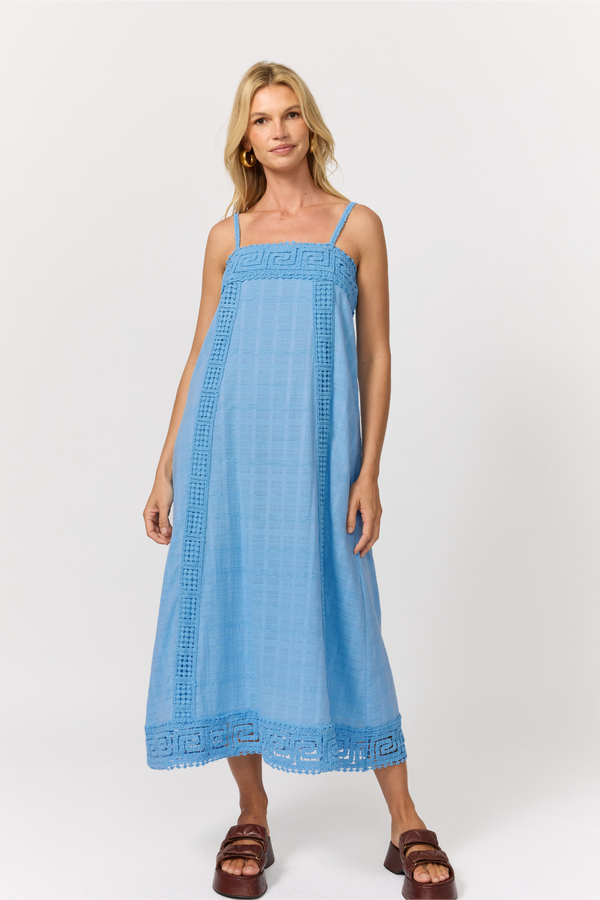 A woman wearing a light blue strappy cotton midi dress with crochet trim at the neckline and hem, paired with brown sandals, against a light gray studio background.