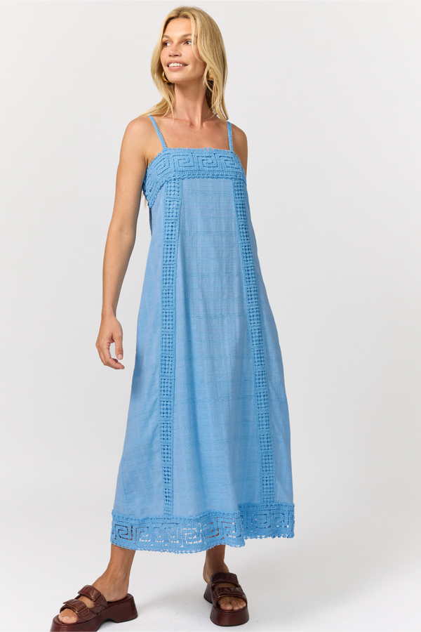 A woman wearing a light blue strappy A-line cotton midi dress with crochet trim panels, shown at a three-quarter angle against a light gray studio background.