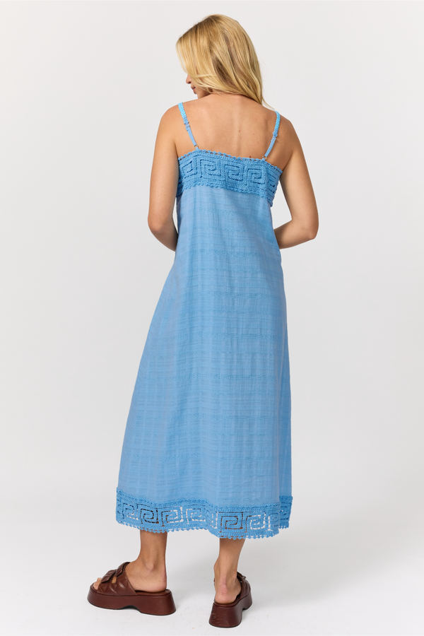 A woman shown from the back wearing a light blue strappy cotton midi dress with a crochet trim hem, paired with brown sandals, against a light gray studio background.