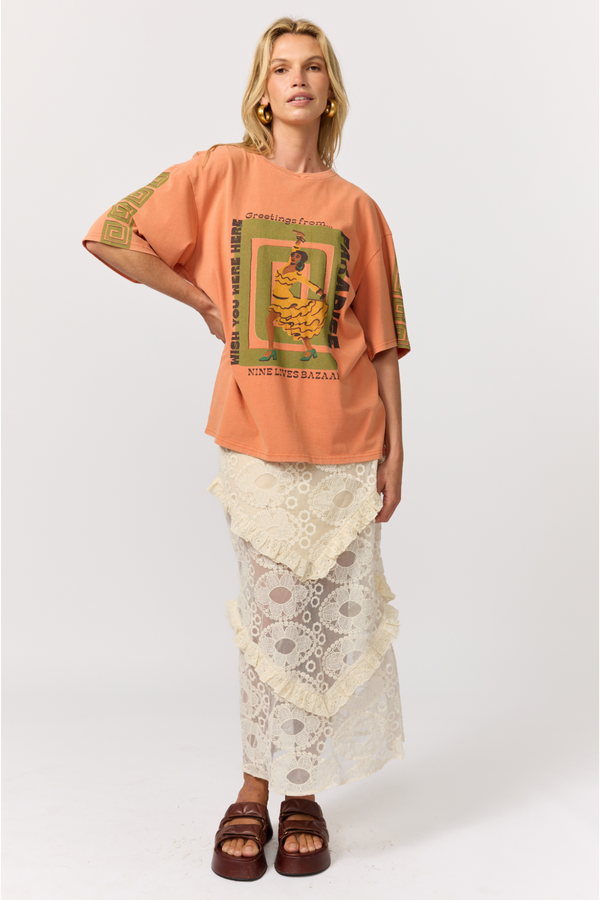 A woman wearing an oversized peach graphic T-shirt with green and yellow print, a cream lace tiered maxi skirt and brown platform sandals, against a light gray studio background.