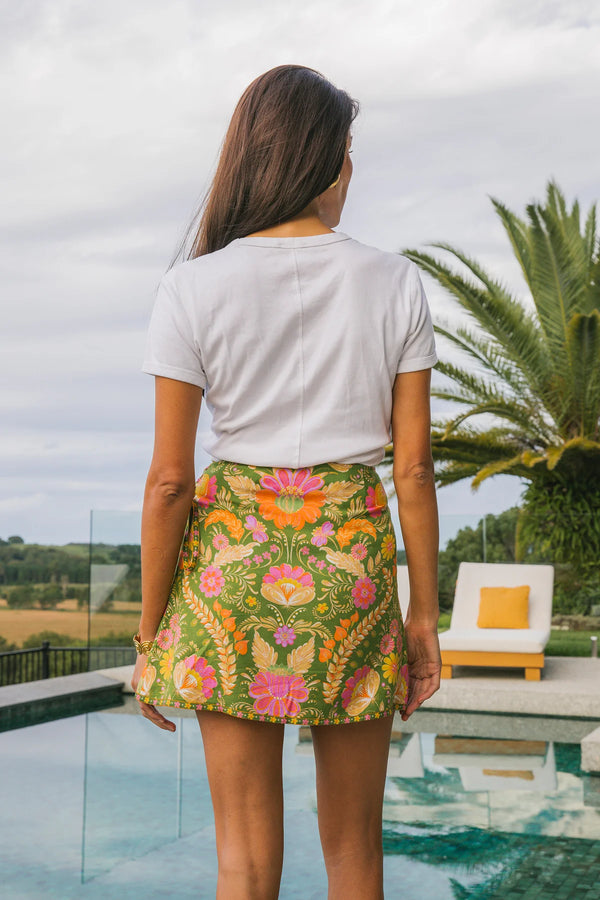 Woman wearing a white t-shirt and colorful floral skirt standing by a pool with palm trees in the background.