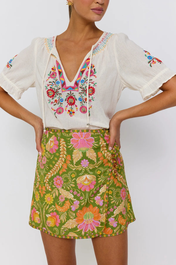 Woman wearing a white blouse with colorful embroidery and a green floral skirt on a white background
