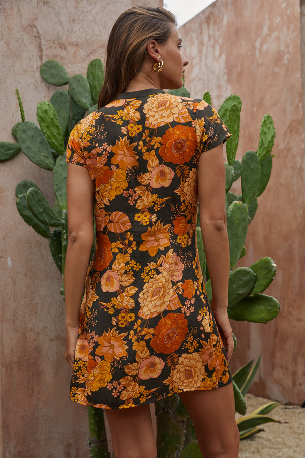 Woman wearing a floral dress with orange and yellow flowers against a textured wall with cacti.