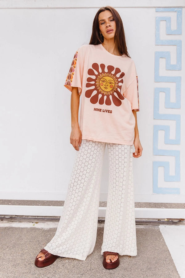 Woman wearing a pink t-shirt with a graphic design and white patterned pants against a white wall.