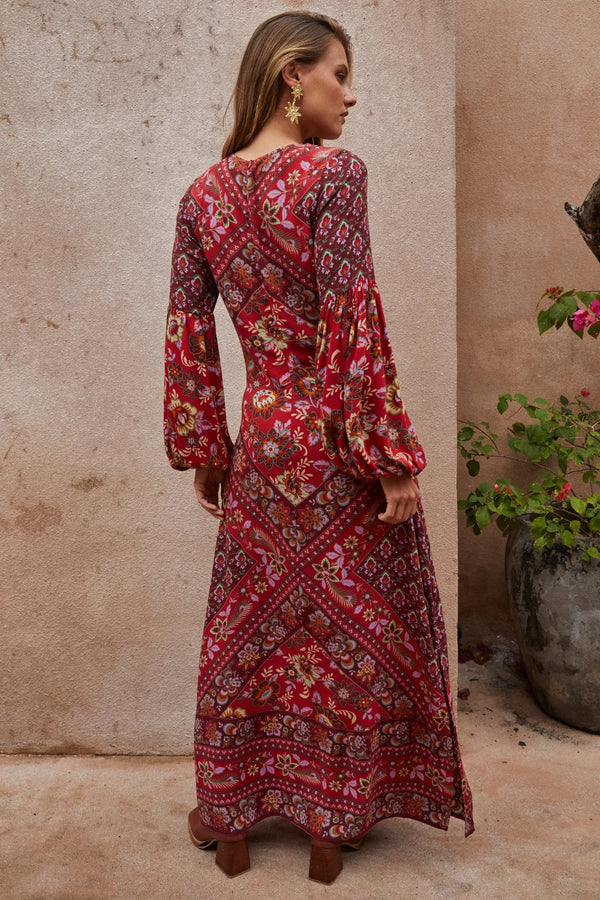 Woman wearing a red patterned dress standing against a beige wall with a plant.