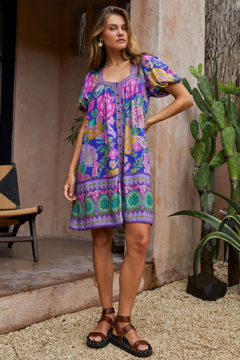 Woman wearing a colorful patterned dress standing outdoors with plants and a chair in the background.