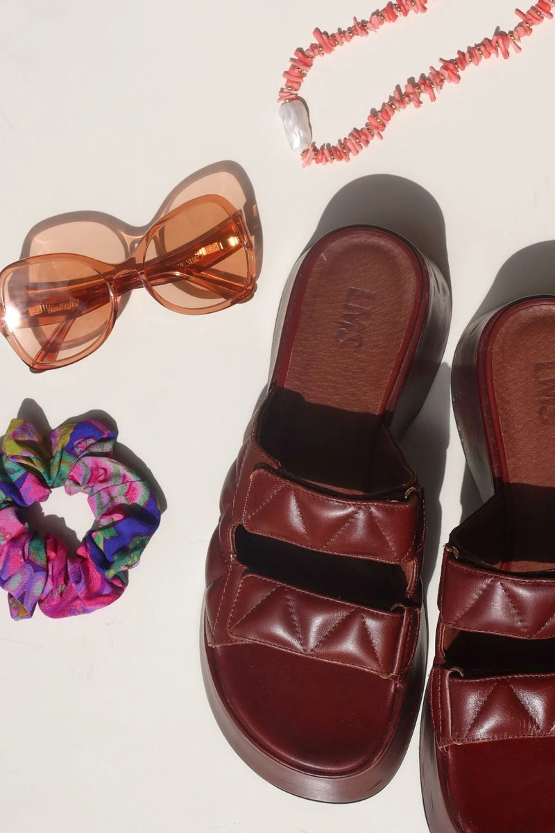 Brown sandals with a colorful scrunchie, sunglasses, and pink necklace on a light background