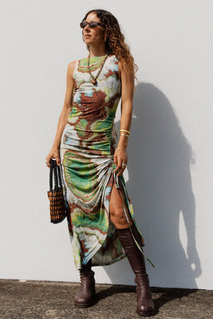 Woman wearing a colorful dress with a slit, paired with brown boots and sunglasses, against a white wall.