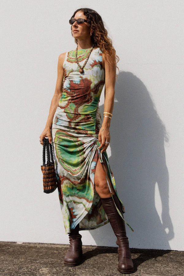 Woman wearing a colorful dress with a slit, paired with brown boots and sunglasses, against a white wall.