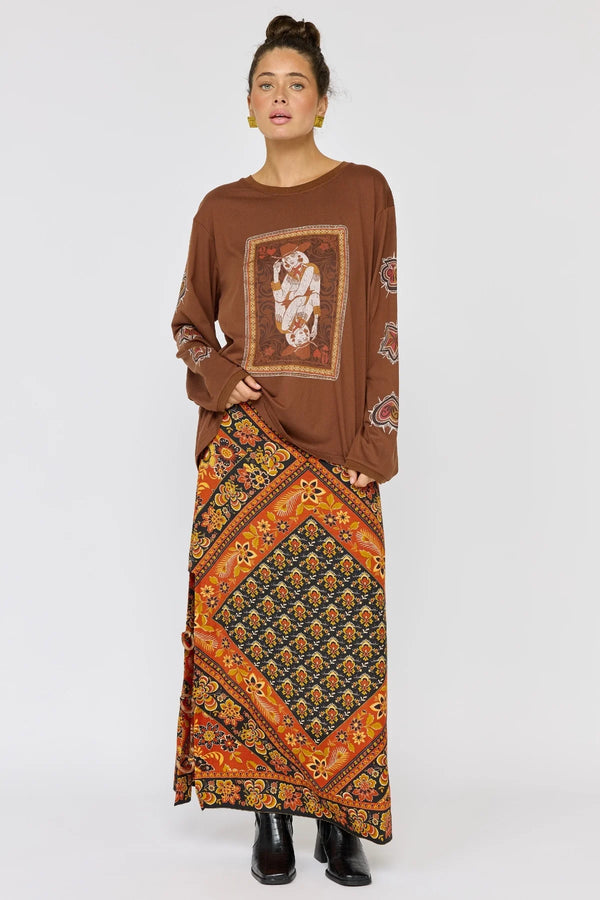 Woman wearing a brown long-sleeve top with a pattern and a long, colorful skirt on a white background