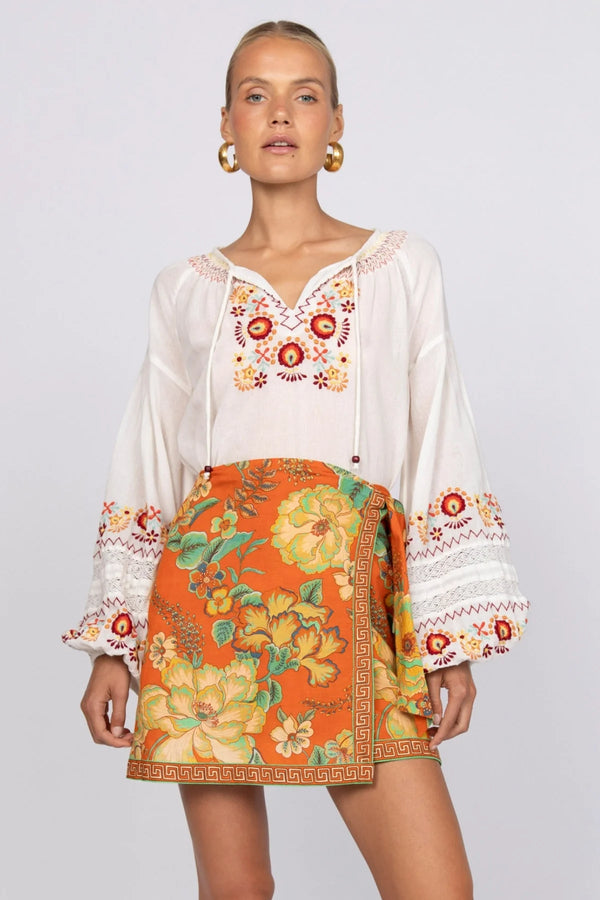 Woman wearing a white blouse with floral embroidery and an orange skirt with floral patterns on a plain background