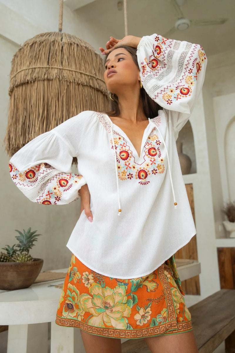 Woman wearing a white blouse with floral embroidery and an orange skirt with floral patterns indoors.