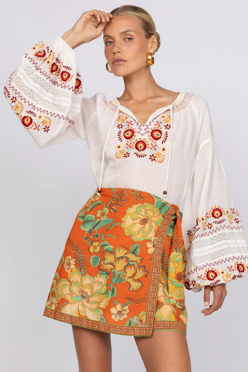 Woman wearing a white blouse with floral embroidery and an orange floral skirt on a light gray background