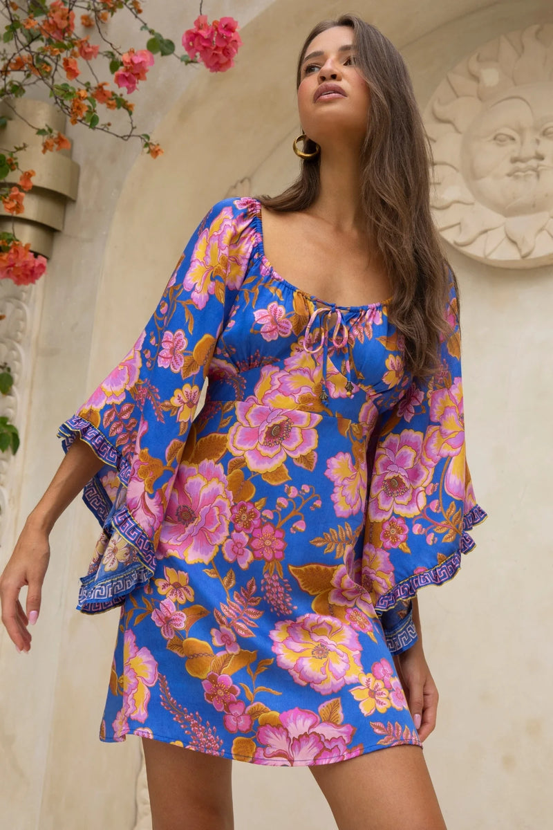 Woman wearing a colorful floral dress standing against an architectural background with flowers.