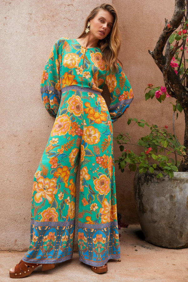Woman wearing a colorful floral dress standing against a beige wall with a plant.