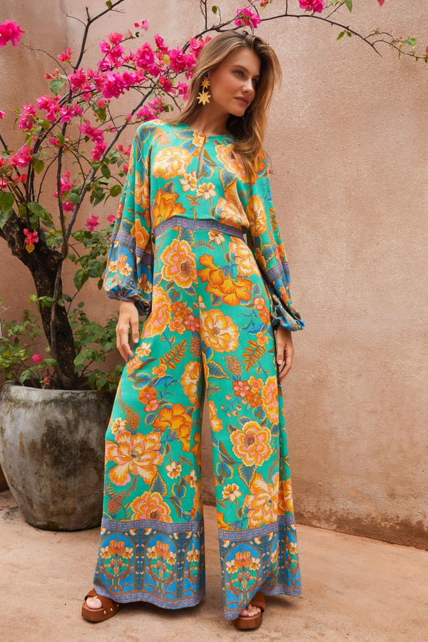 Woman wearing a colorful floral outfit standing next to a potted plant with pink flowers.