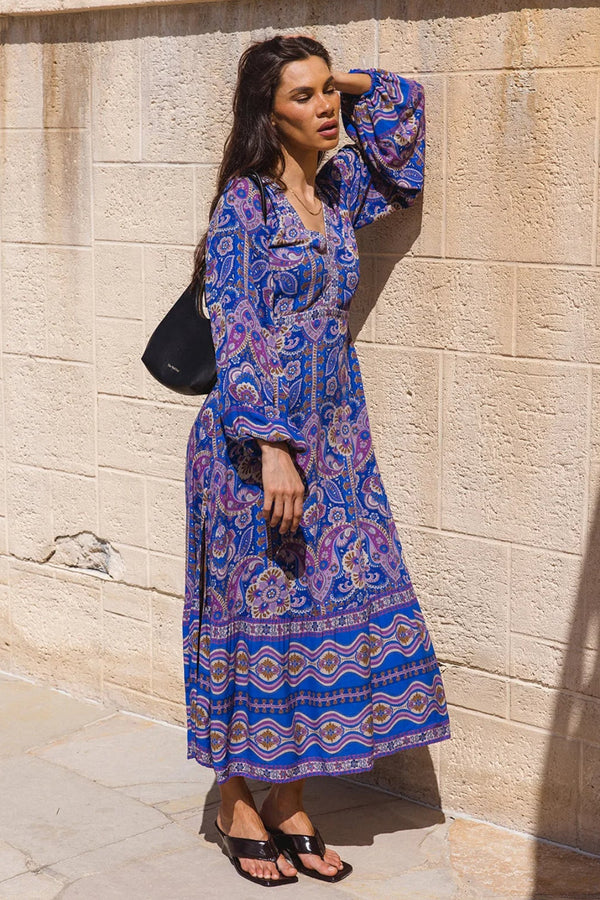 Woman in a blue patterned dress standing against a beige stone wall.