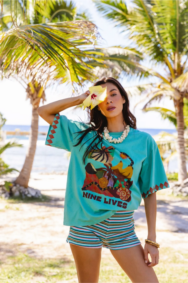 A woman posing on a tropical beach wearing a turquoise graphic t-shirt with 'NINE LIVES' printed on the front, striped shorts, and a white beaded necklace. Location is Cook Islands.