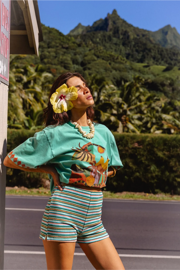 A woman posing outdoors in a vibrant mint green crop top with a colorful tropical print and matching striped shorts, set against a lush tropical mountain background.