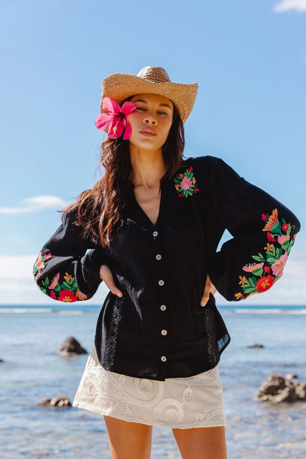 Woman wearing a black embroidered blouse and straw hat with a pink flower on a beach.