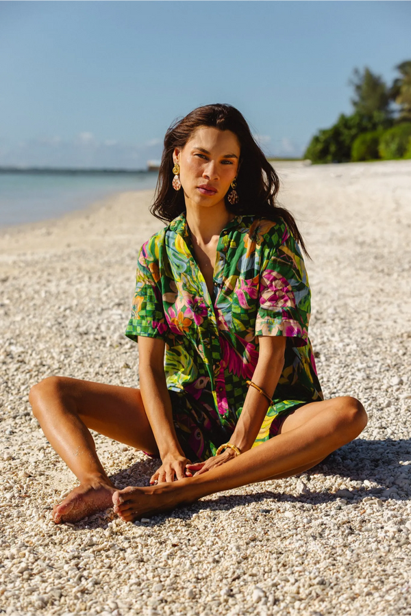 A woman sits cross-legged on a sandy beach wearing a colorful tropical floral print short-sleeve shirt and matching shorts. The background shows a calm blue ocean and sky. Location is Cook Islands.