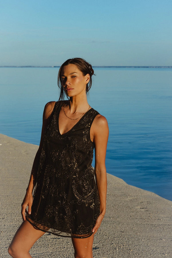 A woman wearing a sleeveless black lace mini dress stands on a sandy shore with blue water and sky in the background.