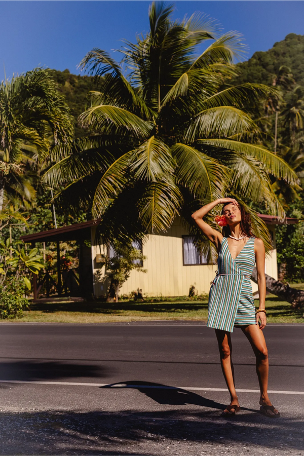 A woman stands on a paved road in front of a tropical resort with palm trees and a small building, wearing a green and white vertically striped halter-neck romper. Location is Cook Islands.