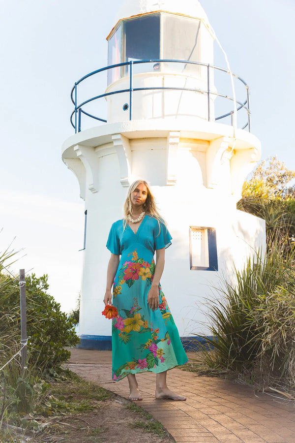A woman wearing a turquoise floral maxi dress stands on a brick path in front of a white lighthouse. The dress features a vibrant multicolor floral pattern with pink, yellow, and orange flowers.