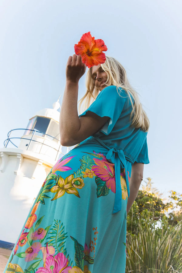 A woman wearing a turquoise blue maxi dress with a vibrant tropical floral print stands outdoors in front of a lighthouse, holding a large red hibiscus flower in front of her face.