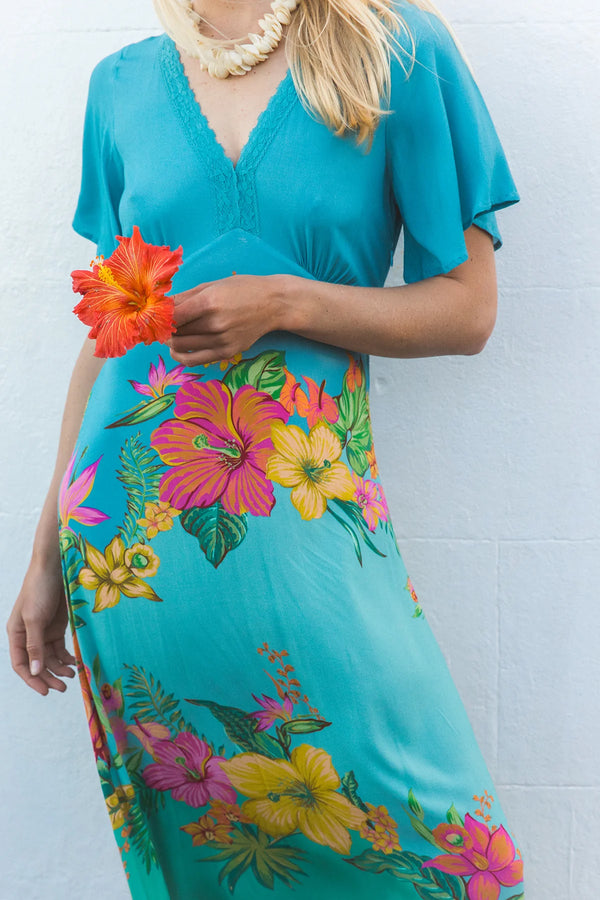 A woman wearing a turquoise maxi dress with a vibrant multicolor floral print, holding a bright orange hibiscus flower against a light gray brick wall.