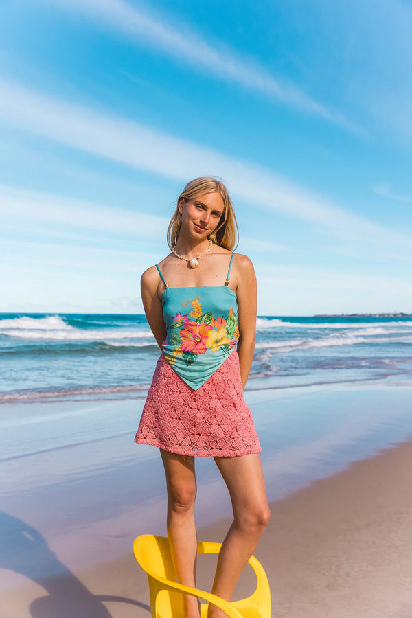 A woman stands on a sandy beach wearing a two-piece swimsuit with a turquoise floral top and a pink crochet skirt, with the ocean and blue sky behind her.