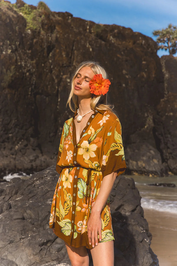 A woman stands on a rocky beach wearing a short brown floral romper with yellow, orange, and green flowers, with an orange flower tucked into her hair. Large dark rock cliffs and a patch of blue sky are in the background. Location Dreamtime Beach, Fingal Head, NSW.