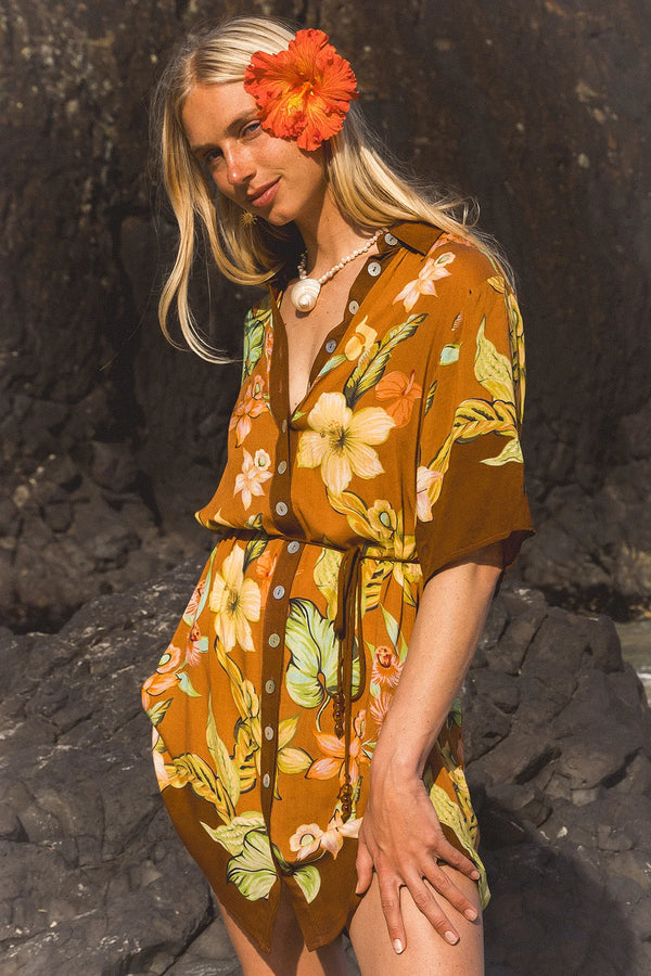 A woman wearing a brown short-sleeve floral romper with yellow, orange, and green botanical print, standing outdoors against a dark rocky background with a large orange flower in her hair. Location Dreamtime Beach, Fingal Head, NSW.