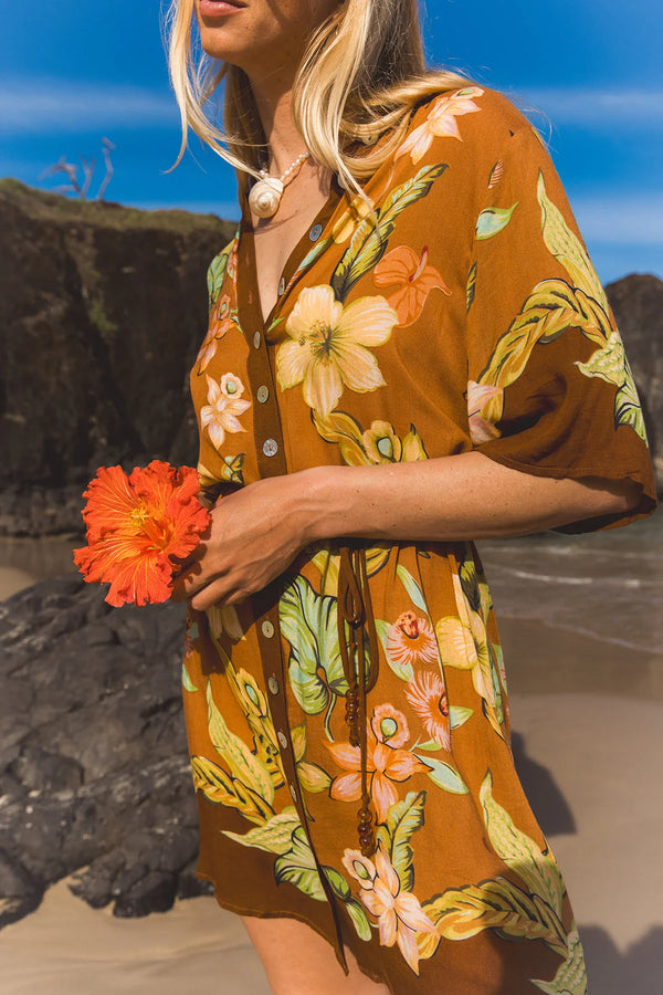 A woman on a beach wearing a brown short-sleeve button-front cover-up dress with a colorful tropical floral print. She is holding a bright orange hibiscus flower, with dark rocks and blue sky in the background.  Location Dreamtime Beach, Fingal Head, NSW.