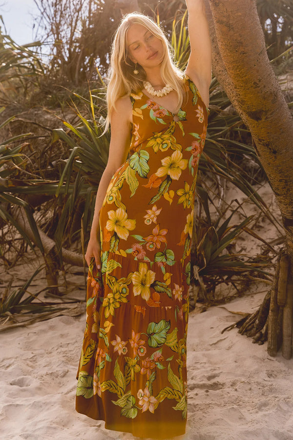 A woman wearing a sleeveless brown maxi dress with a colorful yellow, green, and orange floral print, standing on a sandy beach next to a tree trunk. Location Dreamtime Beach, Fingal Head, NSW.