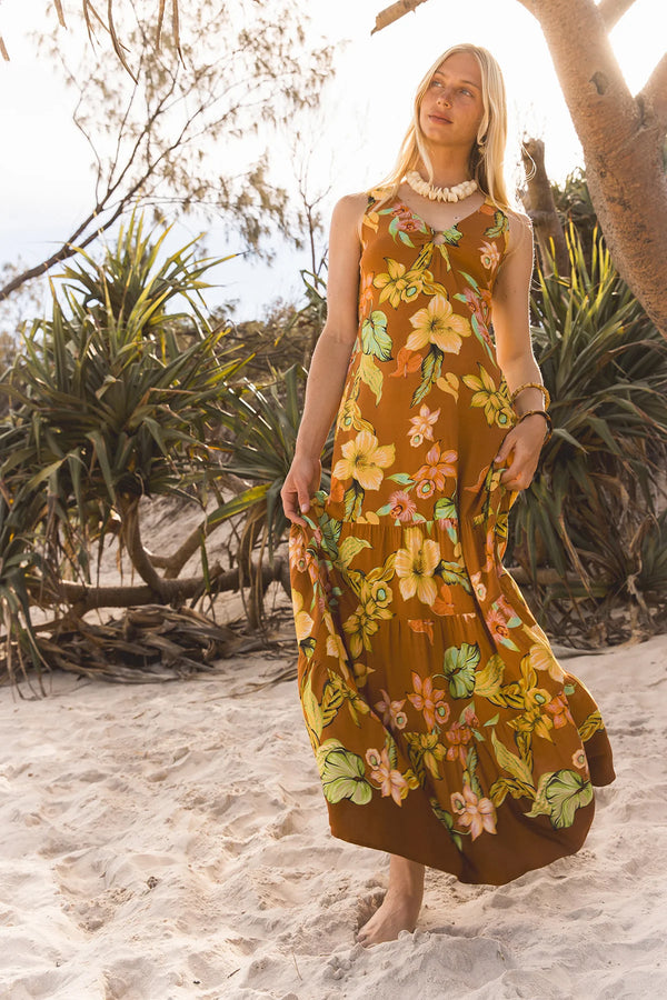 A woman stands barefoot on sandy ground wearing a sleeveless, midi-length maxi dress with a retro floral print in mustard brown, yellow, orange, and green. The background features beach plants, trees, and a bright, sunlit sky.