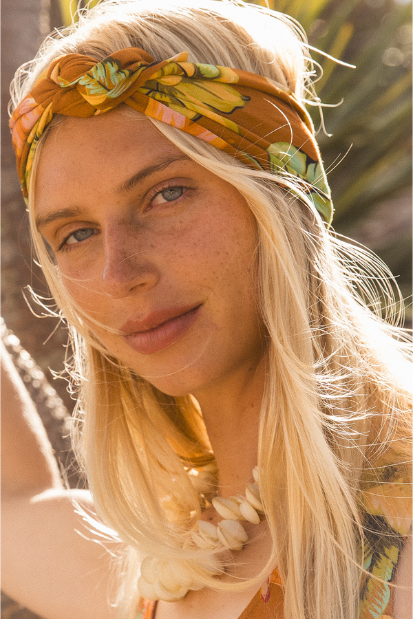 A close-up portrait of a woman with long blonde hair wearing a patterned orange and green headband, a shell necklace, and a matching patterned top, set against a warm, sunlit outdoor background.