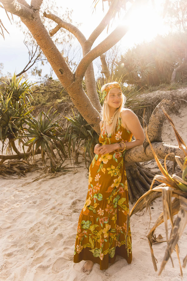 A woman stands on a sandy beach wearing a long sleeveless maxi dress with a yellow, orange, and green floral print. She is posed under the branches of a tree with bright sunlight shining through the background.