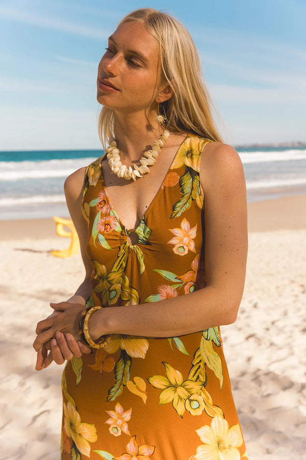 A woman wearing a yellow-orange floral halter dress stands on a sandy beach with the ocean and blue sky in the background. Location Dreamtime Beach, Fingal Head, NSW.