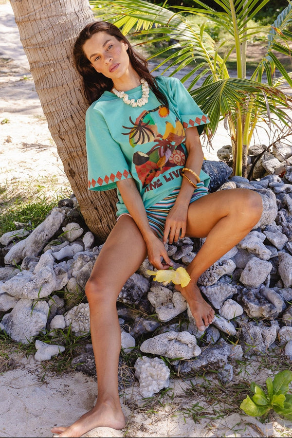 A woman sits barefoot on white rocks beside a palm tree, wearing a turquoise graphic t-shirt with a colorful tropical print and matching patterned shorts in a sunny outdoor tropical setting.
