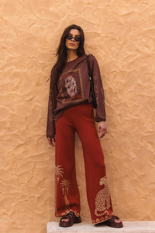 Woman wearing a patterned top and red pants against a textured beige wall.