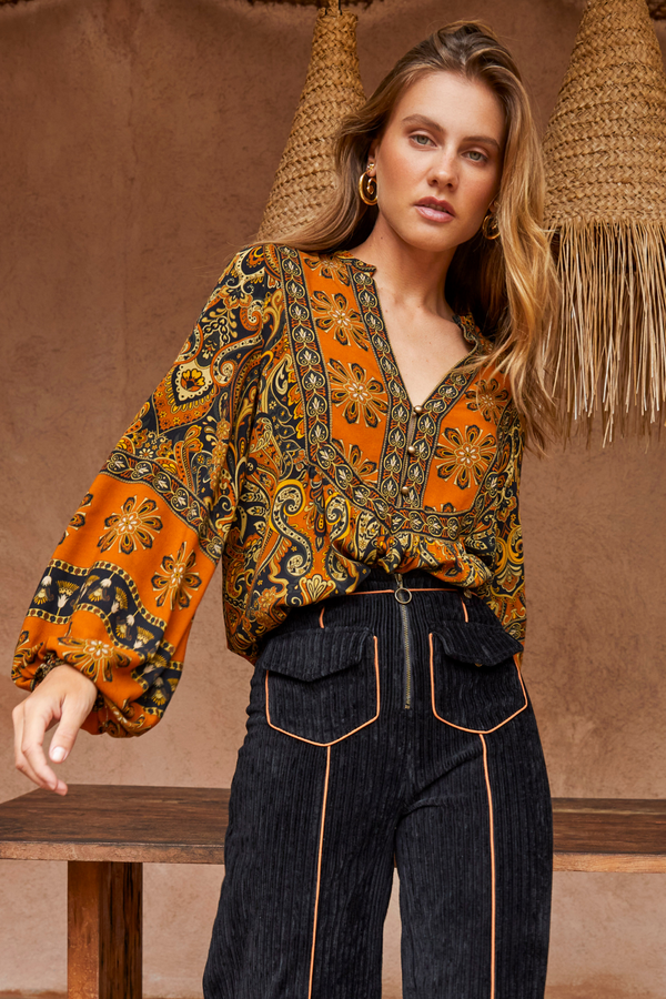 Woman wearing a patterned orange and black blouse with high-waisted dark jeans against a brown background.