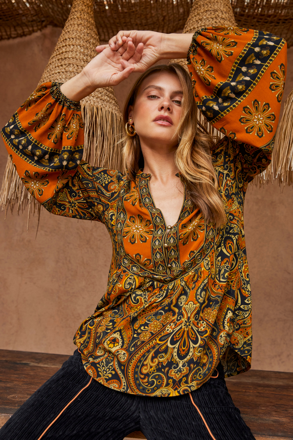 Woman wearing a patterned orange and black top with fringed sleeves against a brown background