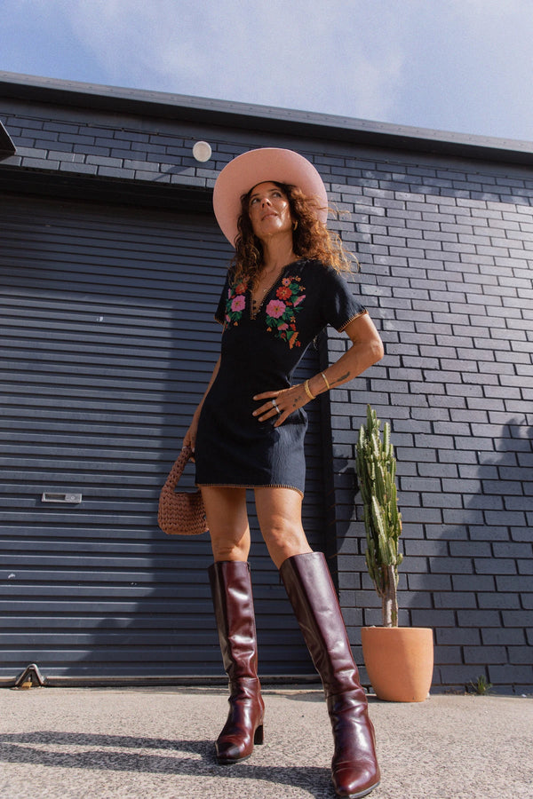 A woman stands outdoors in front of a dark gray brick wall and garage door, wearing a black short-sleeve mini dress with colorful floral embroidery at the neckline, a wide-brimmed light pink hat, dark brown knee-high leather boots, and carrying a woven brown handbag.