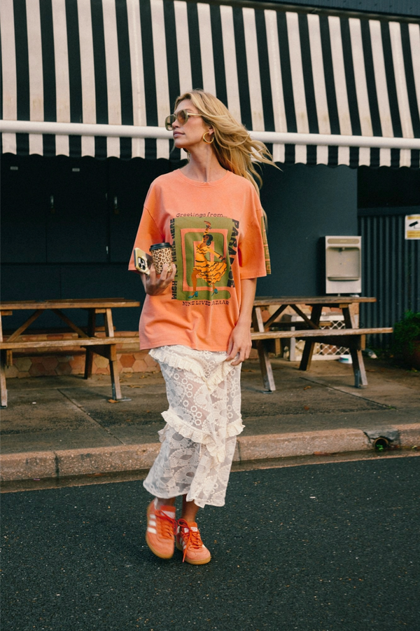 A woman stands outdoors in front of a building with a black-and-white striped awning, wearing an oversized peach graphic T-shirt, white lace ruffled pants, and orange sneakers.