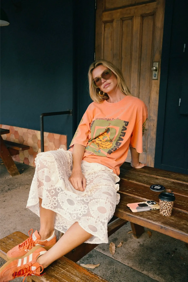 A woman sitting on a wooden bench wearing an oversized peach graphic T-shirt, a cream lace ruffle tiered maxi skirt and orange sneakers, in front of a dark painted wall.